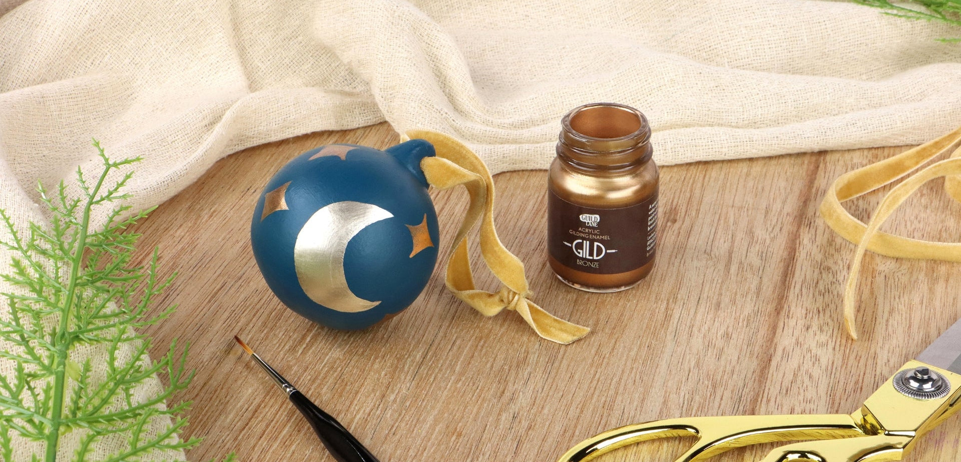 Blue decorative item with white crescent moon and gold star, brown bottle labeled 'GLD', and gold scissors on a wooden surface.