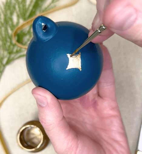 Blue ornament being decorated with gold paint using a small brush.
