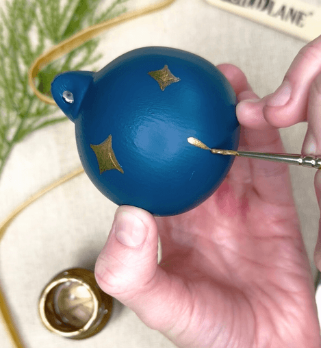 Hand holding a blue decorative ornament with gold accents, using a tool to apply or adjust something on it.