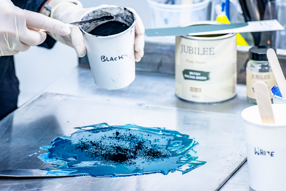 Person mixing black paint with a blue substance on a white surface, with 'Jubilee' products in the background.