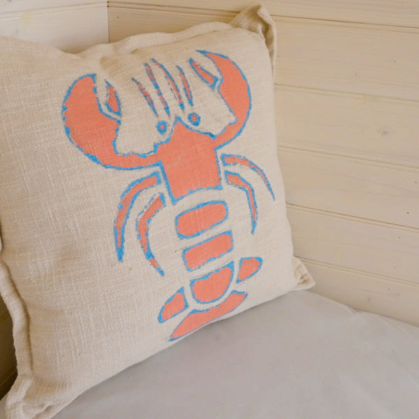A square beige linen throw pillow featuring a stylized lobster design in coral orange and bright blue, resting against a light wood-paneled wall.