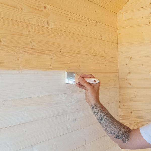 A person's tattooed arm holding a metal paint brush with a white handle, applying a light-colored finish to a wood-paneled wall corner in a room.