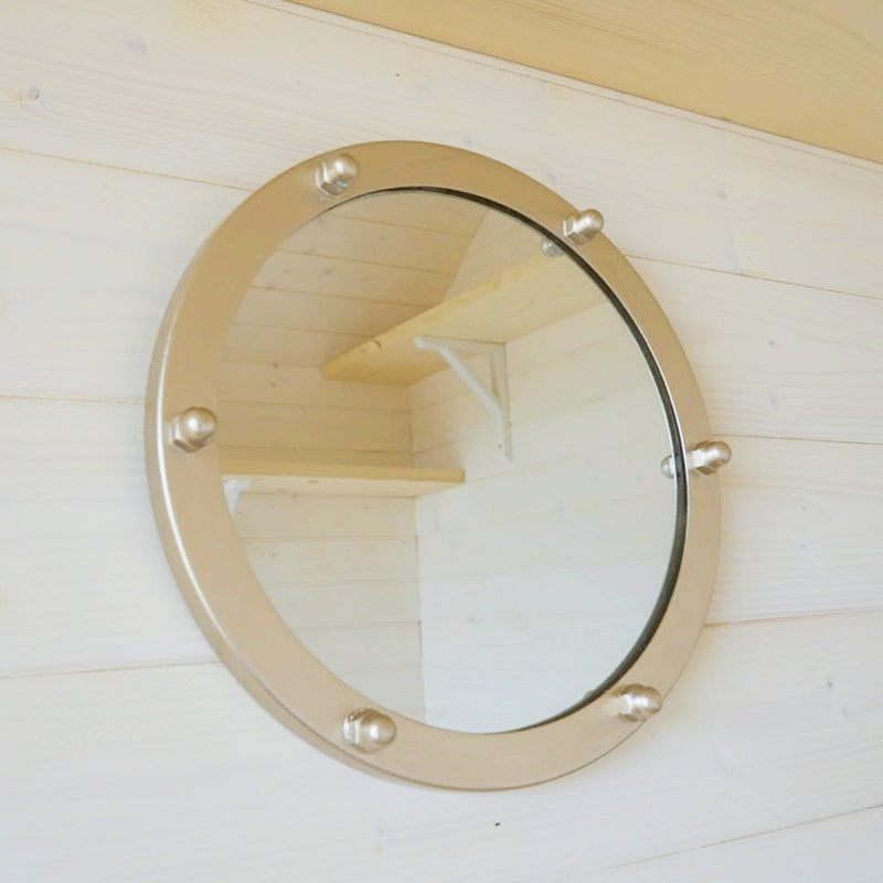 A round wall mirror with a polished brass porthole-style frame and decorative finials, mounted on a light wood-paneled wall.