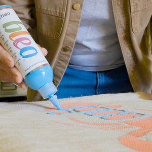
                      
                        A person holding a tube of acrylic paint with a fine tip, applying light blue paint to a canvas with peach-colored lettering.
                      
                    