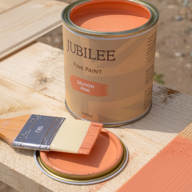 A can of JUBILEE Fine Paint in the shade Salmon Pink, shown outdoors on a wooden surface next to a paintbrush and a paint-dipped metal lid.