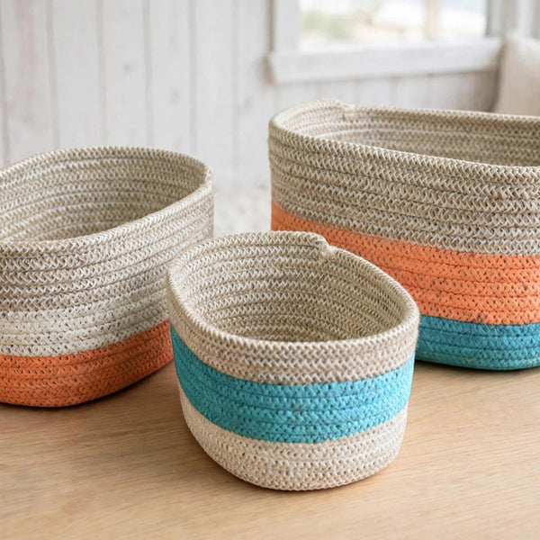 Three woven storage baskets in different sizes on a light wood table, featuring horizontal stripes of natural beige, orange, turquoise blue, and white.