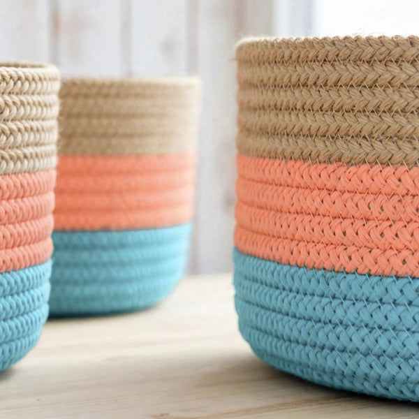 Three woven rope storage baskets with horizontal color bands, shown on a light wood surface with a soft white background.