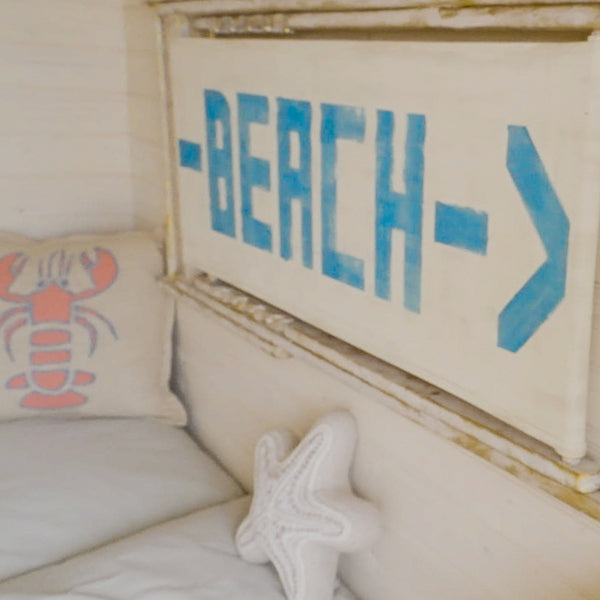 A close-up of a nautical-themed bedroom or playset corner featuring a rustic white canvas sign with blue painted text reading '-BEACH->' and a distressed wooden frame. A white starfish pillow and a beige pillow with a pink lobster graphic are visible on a white bed.