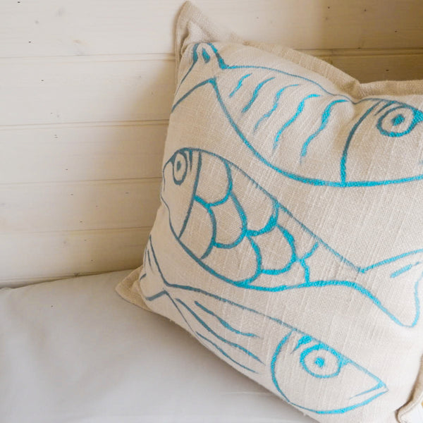 A square throw pillow in a textured cream linen fabric, featuring a hand-drawn style pattern of three fish in bright turquoise blue, resting on a white cushion against a white paneled wooden wall.