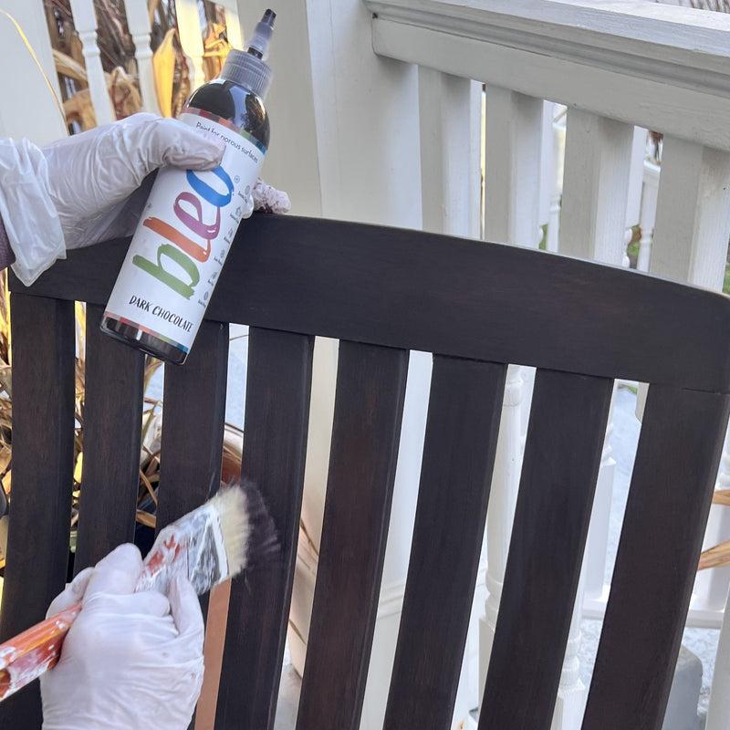 Person applying a dark brown finish to wooden furniture using a bottle labelled 'bleo' and a brush.