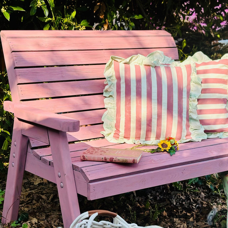 Pink wooden bench with striped cushions and a book in a garden setting