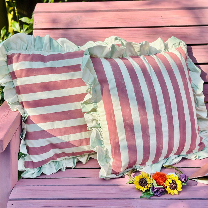 Pink and white striped pillows with ruffles on a pink wooden bench with flowers.