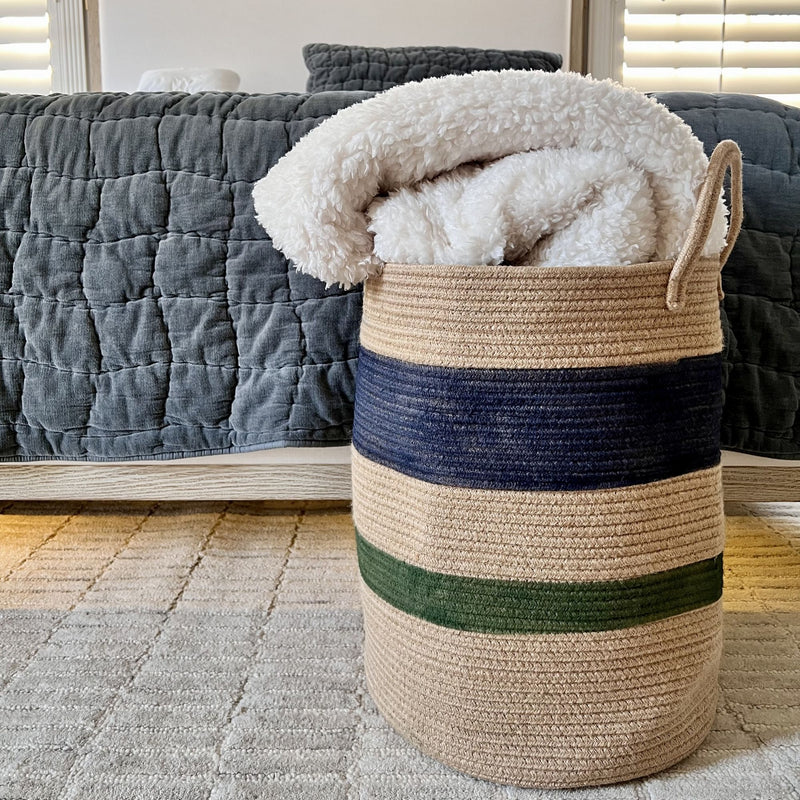 Woven basket with striped design containing a white blanket, placed on a carpeted floor with a couch in the background.