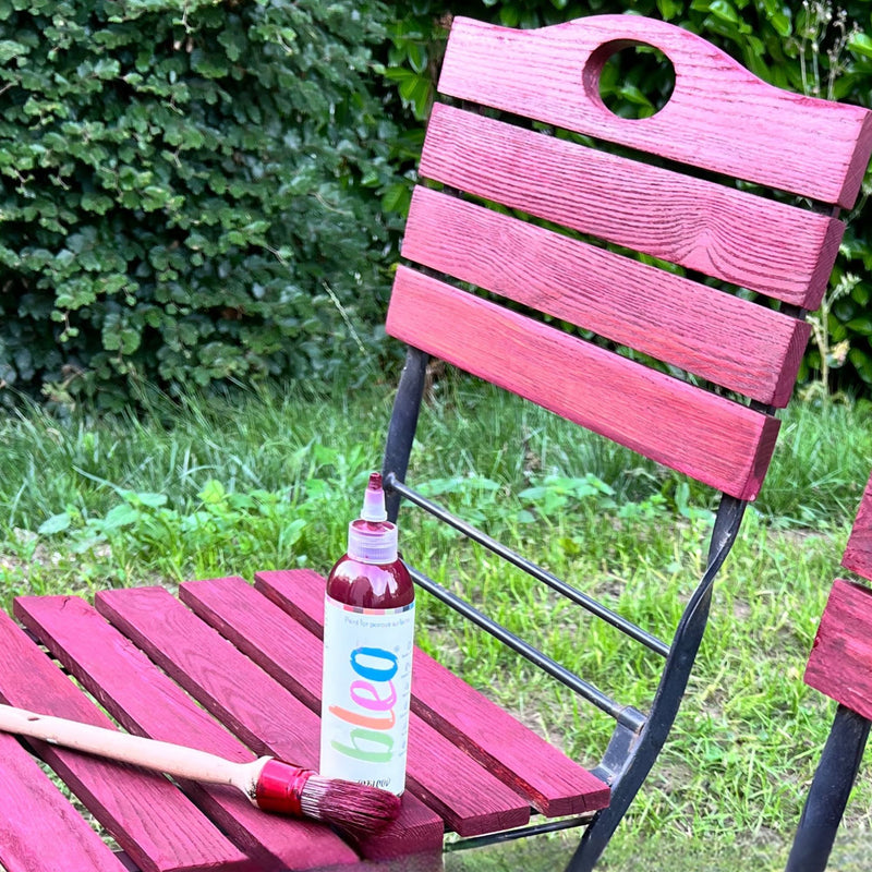 Red wooden chair with a bottle of 'bleo' spray paint and a brush on it, set against a green outdoor background.