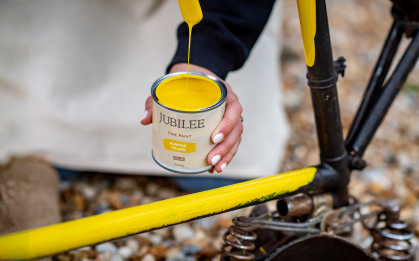 Person holding a can of Jubilee yellow paint next to a bicycle chain on a natural background