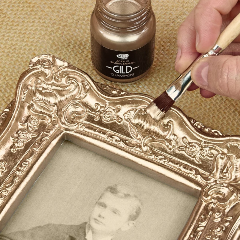 Person gilding an ornate picture frame with champagne-colored gold leaf using a brush.