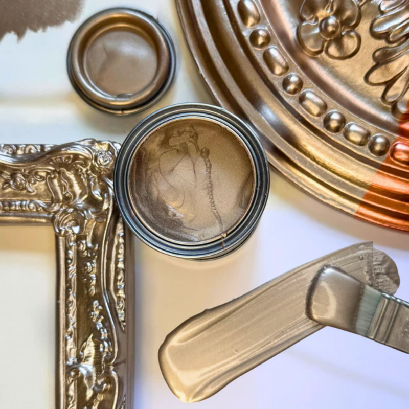 Decorative gold paint can, brush, and ornate frame on a white surface