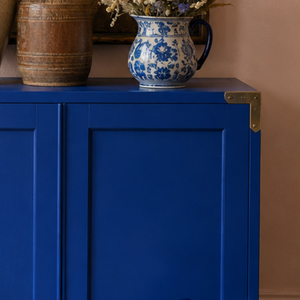 
                      
                        A royal blue wooden cabinet with brass corner brackets, styled with a blue and white ceramic pitcher and a rustic brown vase against a warm beige wall.
                      
                    