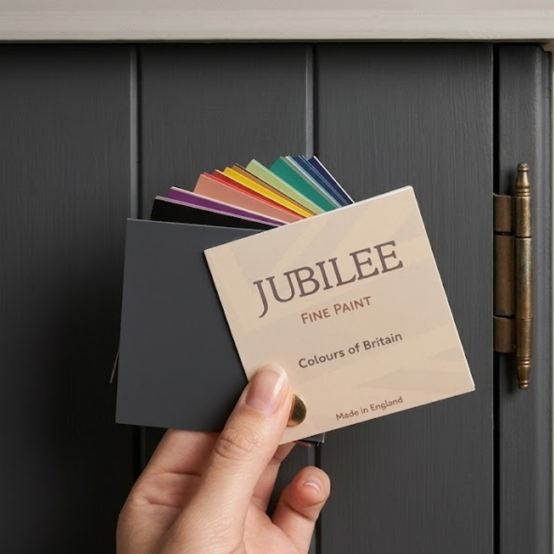 A hand holds a Jubilee Fine Paint 'Colours of Britain' card set in front of a dark grey painted wooden door. The set includes a dark grey card, a beige card, and a fan of various coloured paint swatches.