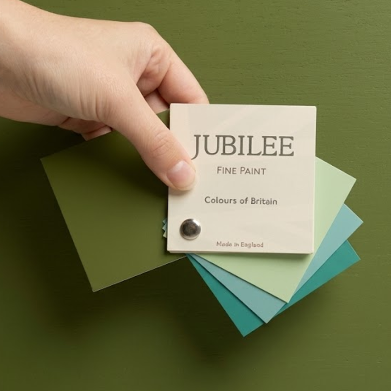 A hand holds a small card labeled JUBILEE FINE PAINT, showing a fanned stack of paint color swatches in olive green, pale green, blue, and teal against a matching olive green background.