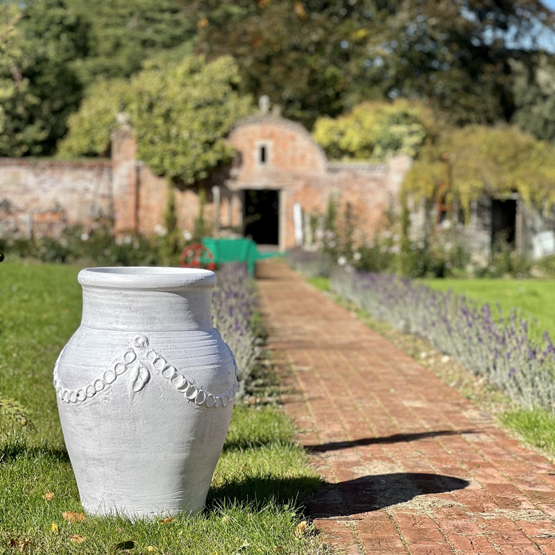 White decorative pot on a grassy path with a garden and brick wall in the background