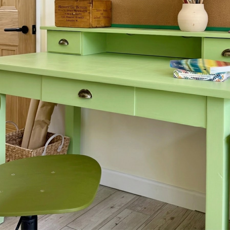 A light green writing desk with a matching green chair, shown in a home office setting with a wooden door, basket, and books on the desk.