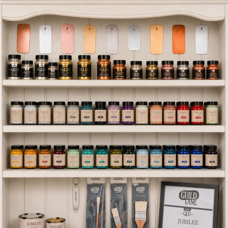 A white wooden display cabinet neatly organized with art supplies from the brand Guild Lane, featuring hanging metallic swatches, a shelf of metallic "Super -GILD-" paint jars, two shelves of brightly coloured "Jubilee" paint jars arranged in a spectrum, and hanging paintbrushes above larger paint tins and a branded sign.