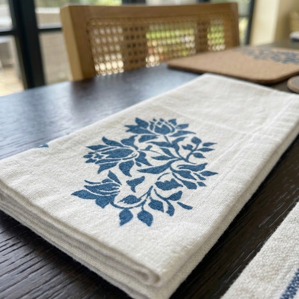 A folded off-white linen napkin with a blue floral block print, displayed on a dark wooden table.