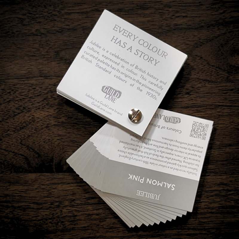 A stack of printed cards or booklets on a dark wooden surface. The top card is white with black text reading 'EVERY COLOUR HAS A STORY' and the brand name 'GUILD LANE'.