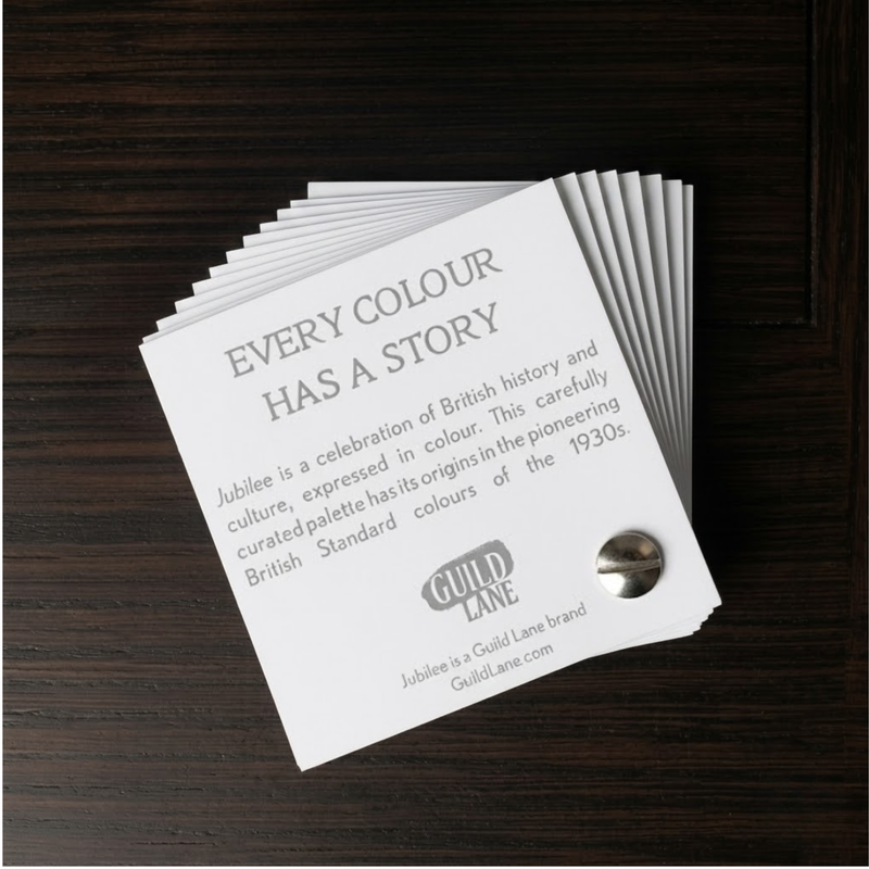 A stack of white printed cards titled 'EVERY COLOUR HAS A STORY' laid on a dark wood surface. The cards feature grey text and a small round metallic badge on the front.