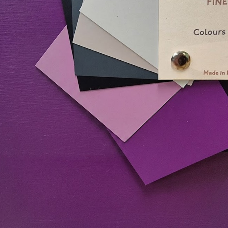 A stack of flat colour sample cards in shades of purple, pink, black, grey, and off-white, arranged on a purple surface with part of a paper label visible that reads 'FINE PAINT'.