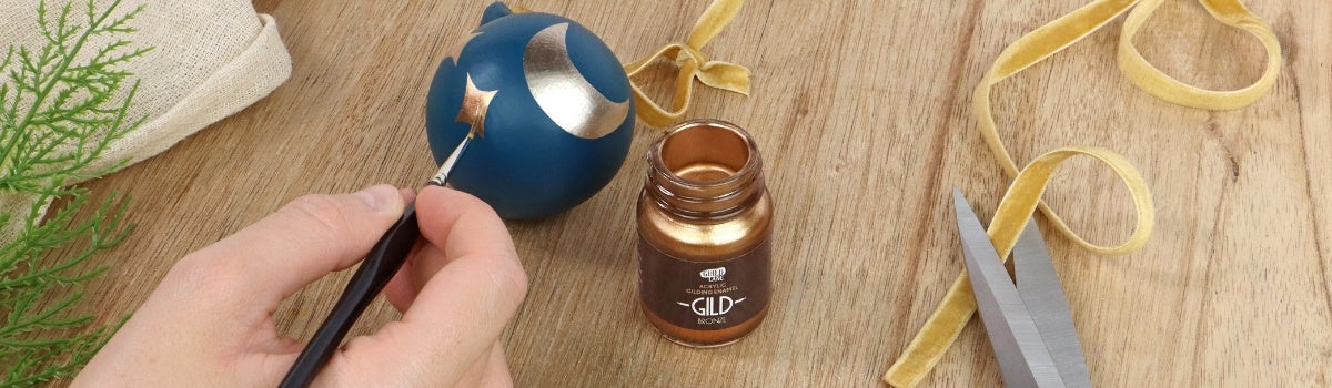 Hand holding a paintbrush near a jar of gold paint, with decorative elements on a wooden surface, painting a bauble