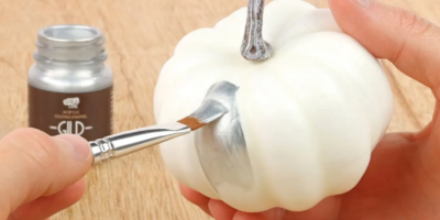Person painting a small white pumpkin with silver using a brush on a wooden surface