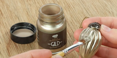 Person applying gold paint to a small decorative item with a brush on a wooden surface.