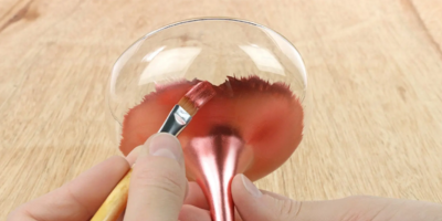 Person using a brush to apply rose gold metallic paint on a transparent glass on a wooden surface