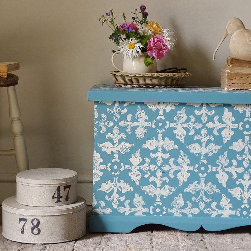 A painted wooden storage chest or bench with a turquoise blue and white damask pattern, styled in a home setting with a basket of flowers on top and vintage boxes nearby.