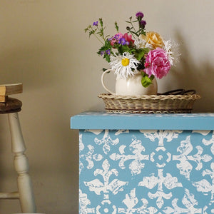 
                      
                        A decorative blue and white patterned storage box or chest with a light blue top, styled with a wicker basket, a white pitcher, and a colorful bouquet of flowers against a beige wall.
                      
                    
