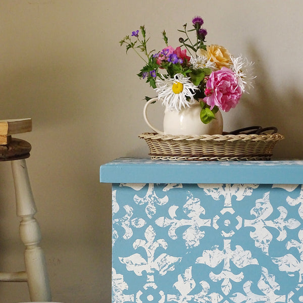 A decorative blue and white patterned storage box or chest with a light blue top, styled with a wicker basket, a white pitcher, and a colorful bouquet of flowers against a beige wall.