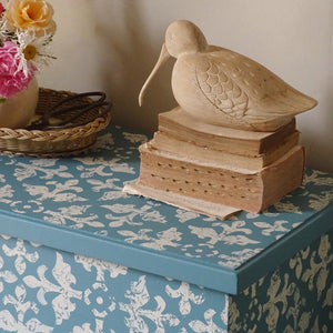 
                      
                        A carved wooden duck figurine perched on stacked vintage books, placed on top of a blue and white patterned storage box in a styled interior setting.
                      
                    