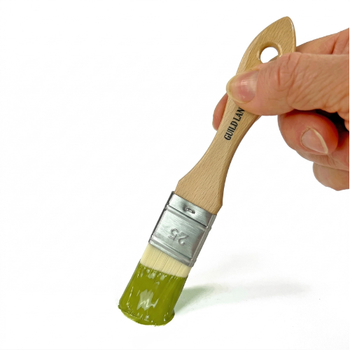 A hand holds a wooden paintbrush. The lower third of the cream-colored bristles is neatly dipped in thick, glossy olive-green paint