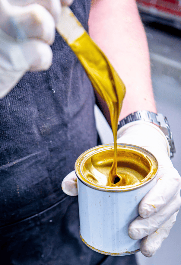 Person pouring metallic super gild gold paint from a can into another container, wearing gloves.