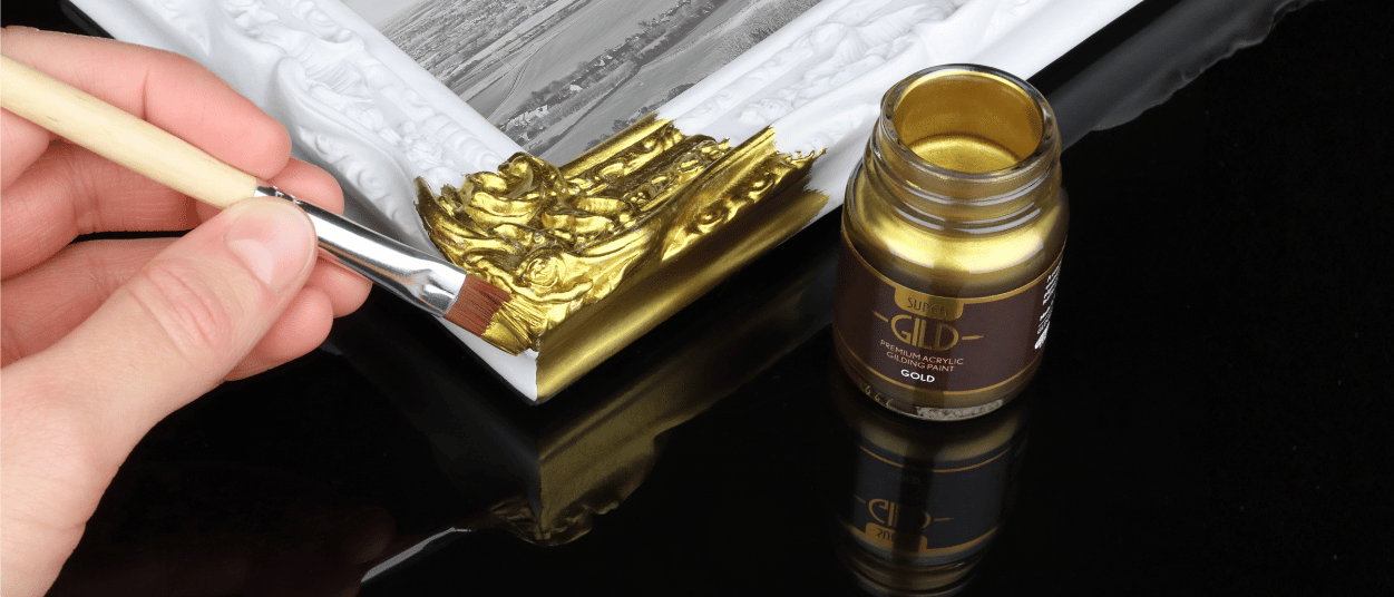 Hand applying gold metallic paint to a decorative frame with a bottle of gold paint in the background.