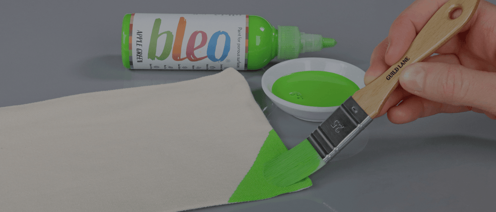 Person painting a canvas with green paint using a brush, with 'bleo' fabric paint bottle and container in the background.