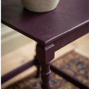 
                      
                        A close-up of a dark plum purple wooden table corner with a turned leg and a woven beige planter on top, set against a blurred indoor background.
                      
                    