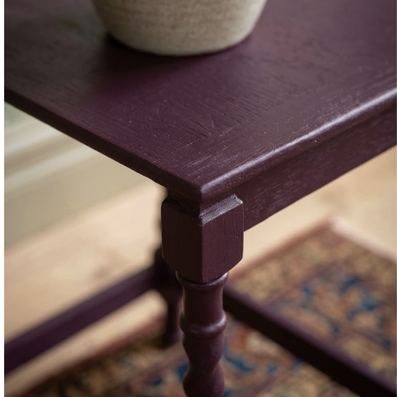 A close-up of a dark plum purple wooden table corner with a turned leg and a woven beige planter on top, set against a blurred indoor background.