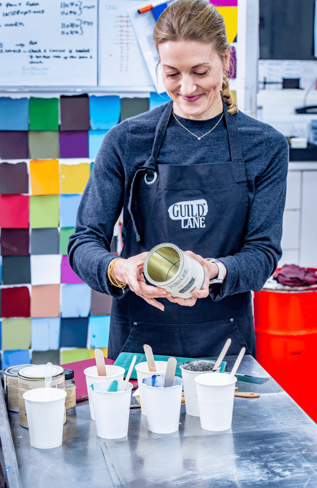 Founder Jeni wearing a 'GUILD LANE' apron holding a container in a workshop setting with colourful materials in the background.