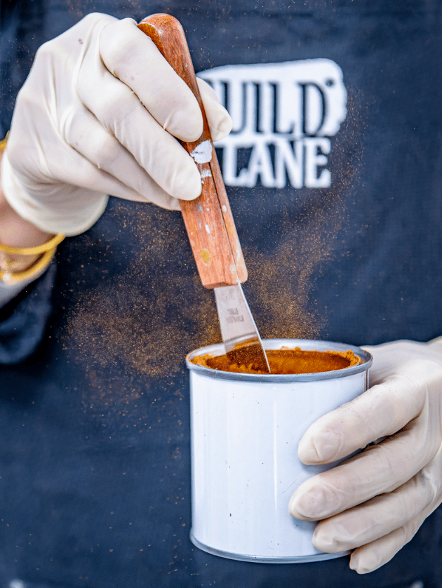 Person wearing gloves using a paint scraper to apply orange paint from a can, with 'GUILD LANE' branding in the background.