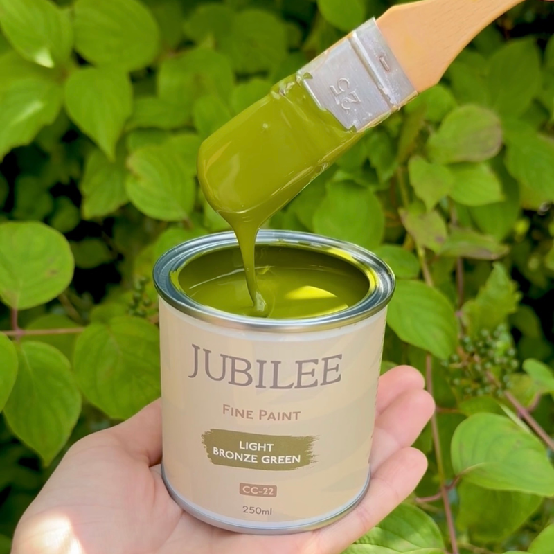 Can of Jubilee Fine Paint with a brush dipping into it, surrounded by green leaves.
