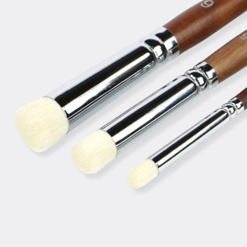 Three makeup brushes with brown wooden handles, shiny silver ferrules, and soft cream-colored bristles, arranged diagonally on a light gray background.