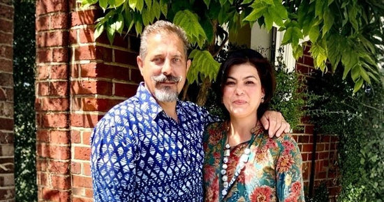 Two people standing together outdoors with a brick wall and greenery in the background